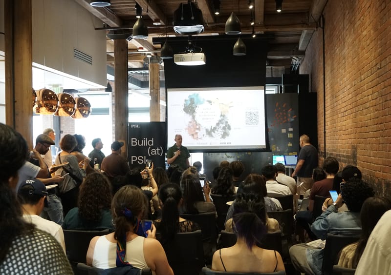 A packed room at Pioneer Square Labs watching a talk, seated audience under warehouse lighting and exposed brick.