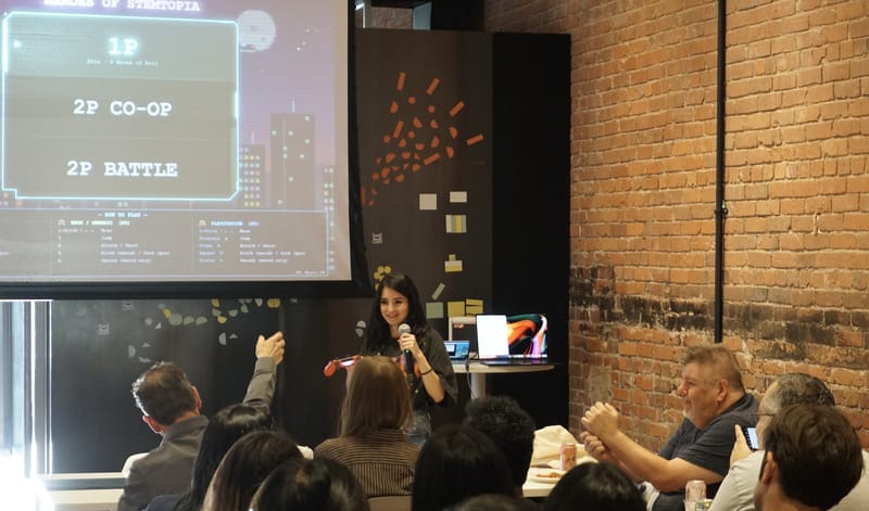A speaker with a microphone in front of a projected Stemtopia game menu showing one player, two player co-op, and two player battle.
