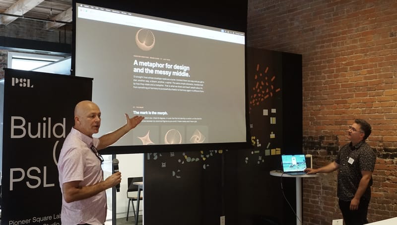 A speaker gesturing at a projected slide titled a metaphor for design and the messy middle while a second presenter stands by the laptop.