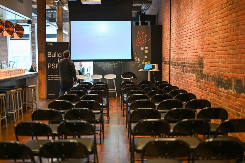 Rows of empty black chairs facing a projection screen in the Pioneer Square Labs event space, under exposed brick and copper pendant lights.