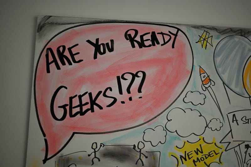 A whiteboard drawing of a speech bubble reading are you ready geeks, surrounded by hand-drawn rockets, clouds and stick figures.