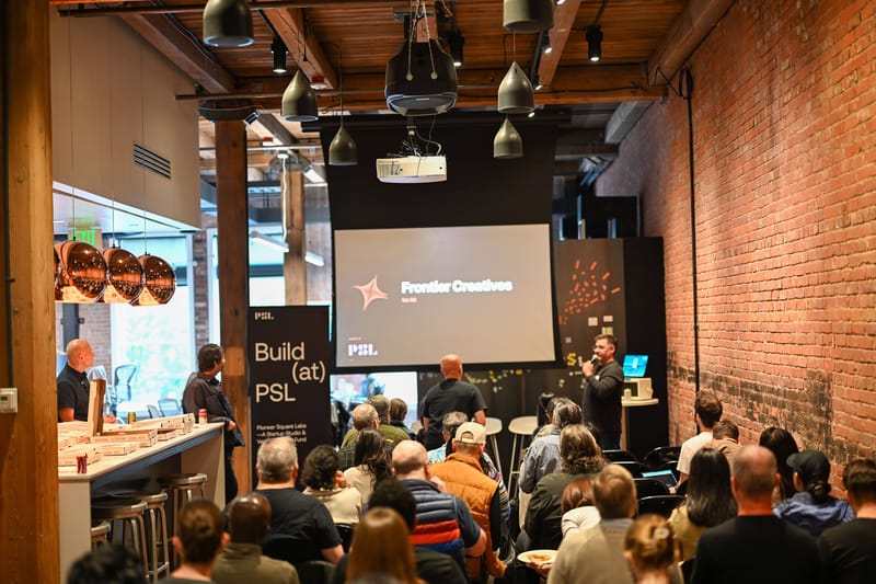 A packed audience seen from the back of the room, facing a projected Frontier Creatives title slide under exposed brick and warehouse lighting.