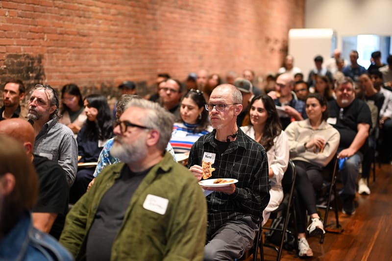 Attendees seated and standing along the side of the room, some holding plates, watching a talk.