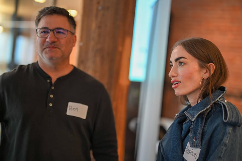 Two attendees in handwritten name tags talking beside a brick column.