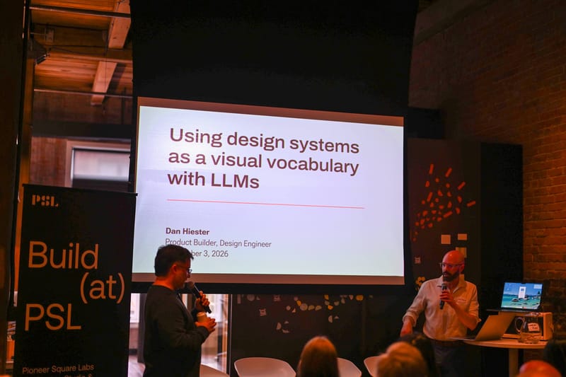 A presenter with a microphone beside a projected slide reading using design systems as a visual vocabulary with LLMs.