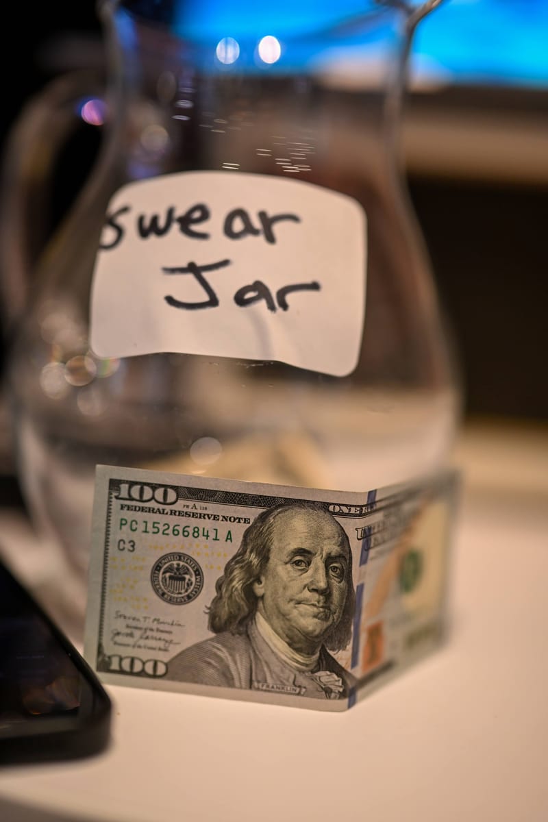 A glass jar labelled swear jar in handwriting with a hundred dollar bill inside.
