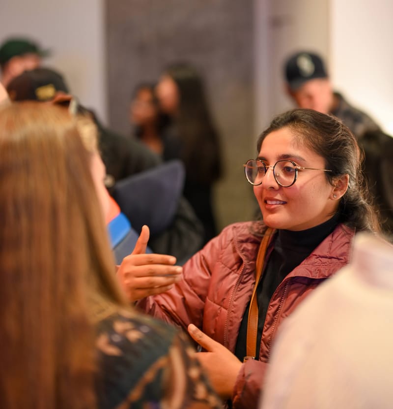 An attendee in a pink jacket gesturing while talking with someone in the foreground.