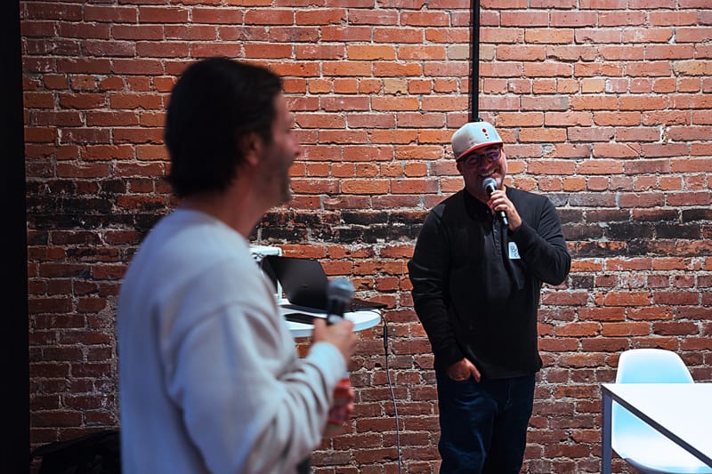 Two hosts with microphones talking in front of an exposed brick wall, one in a white sweatshirt, one in a cap.