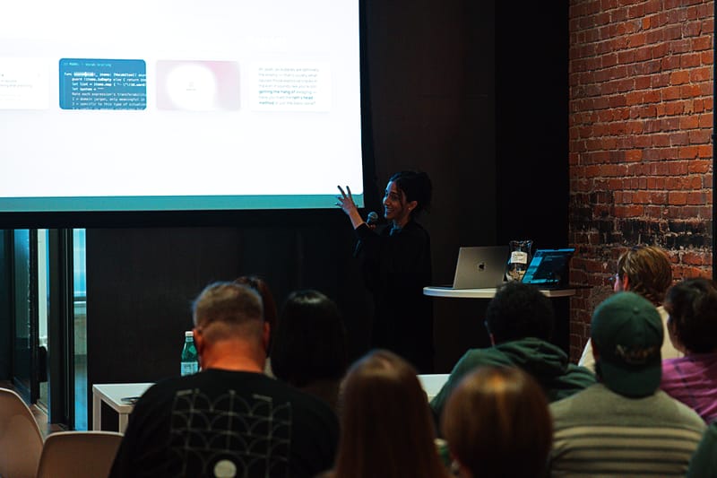 A presenter gesturing beside a projected slide of code and UI mockups, a laptop on a small table, seen over a seated audience.