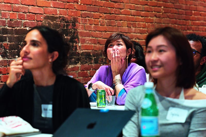 Three attendees in the front row reacting to a talk, one with a hand over her mouth, a laptop and sparkling water on the table.