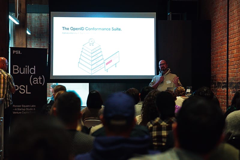 A presenter at a lectern beside a projected slide reading The OpenID Conformance Suite, over the heads of a seated audience.