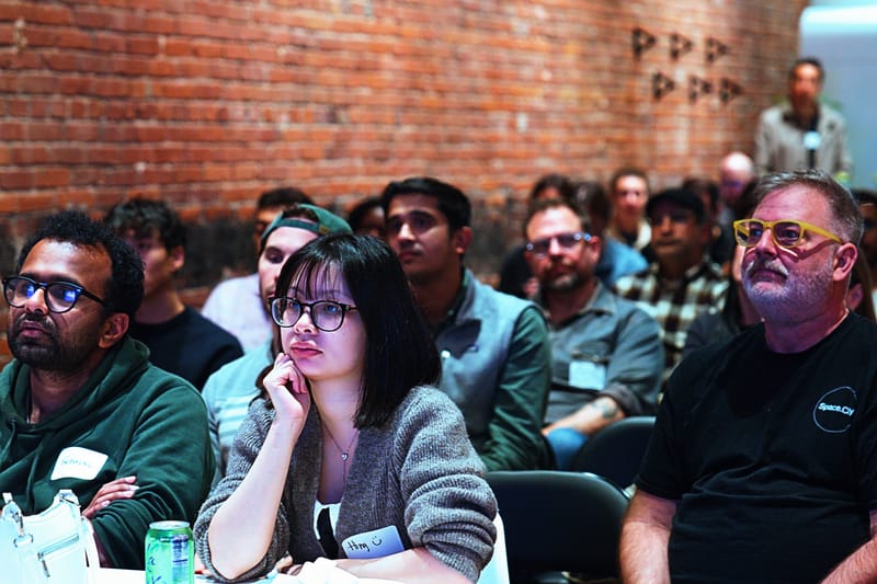 A seated crowd watching a talk, faces lit from the front, brick wall behind.