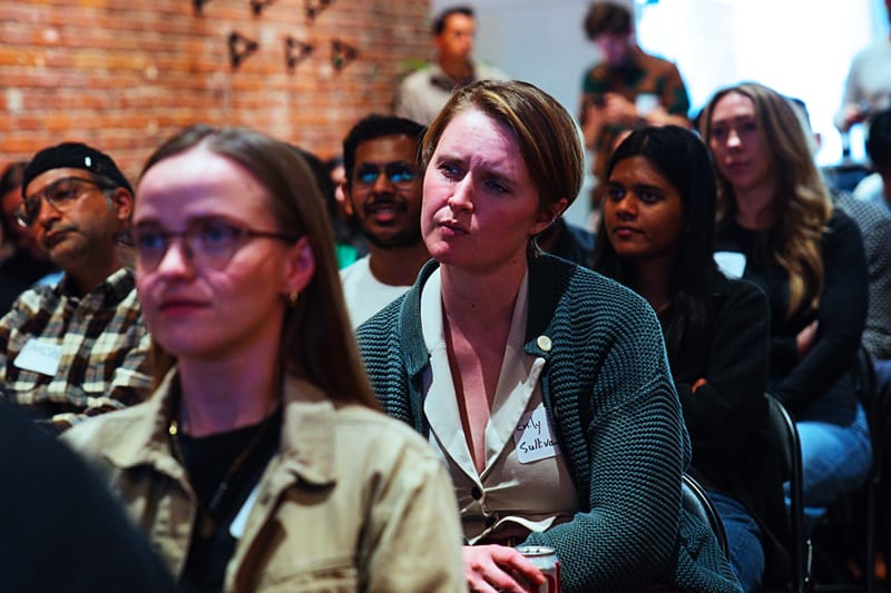 Two attendees in the audience listening closely, one with glasses, others out of focus behind them.