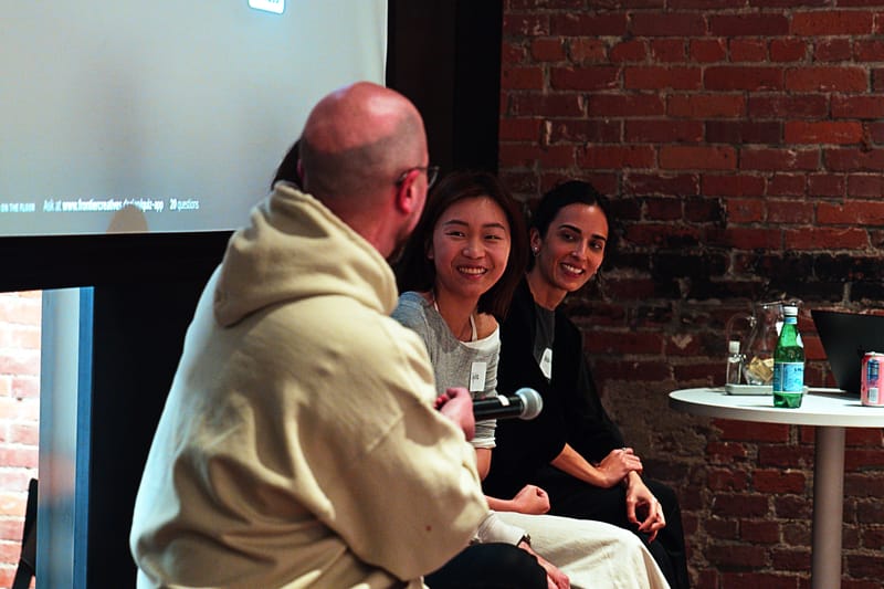 Two panelists laughing while a third, seen from behind in a hoodie, speaks into a microphone.